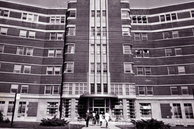 MGH Archive Photo of Coxwell Entrance Black and white archival photo of Toronto East General Hospital entrance with vertical brick columns and curved wings extending on either side. Several people are walking near the entrance.