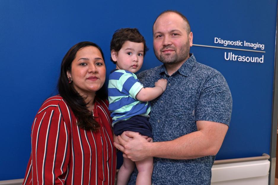 A husband and wife stand with a baby in their arms in front of a blue wall.
